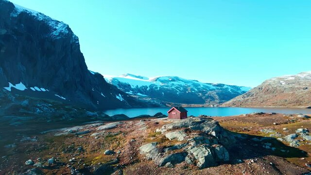 Small hut in the high mountains with a steep tall mountain slope wall and a lake in the background. 4K aerial video.