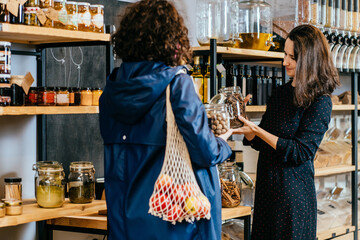 Shop assistant helping customer in bulk food store. Seller advising woman in her purchase of groceries without plastic packaging in zero waste shop. Sustainable shopping at small local businesses. - obrazy, fototapety, plakaty