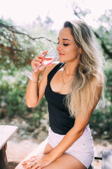 Fototapeta premium Beautiful girl in a juniper and pine forest drinking wine or champagne sitting on a wooden pallet, during the day