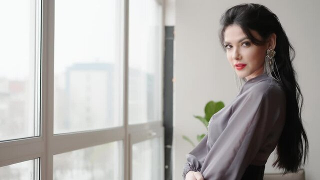 Woman's portrait from the back. A young woman with a beautiful Italian appearance, walks near the window and looks seductively into the camera. She shows off her makeup and flirts