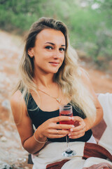 A beautiful girl in a juniper and pine forest with a glass of champagne or wine sits on a wooden pallet, during the day