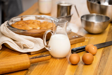 Tasty apple pie on a oaken wood table with a ingredients. Woman in a gray pajama cooking a delicious apple pie on a kitchen in a loft style. Milk, eggs, sugar and a baked pie on a table. Home kitchen.