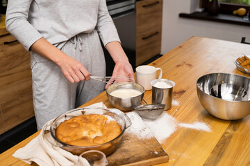 Tasty apple pie on a oaken wood table with a ingredients. Woman in a gray pajama cooking a delicious apple pie on a kitchen in a loft style. Milk, eggs, sugar and a baked pie on a table. Home kitchen.