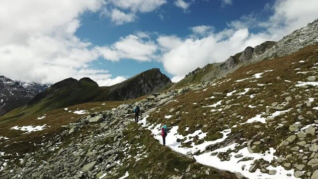 A couple with big backpack hiking in the Hohe Tauern region in Austria. The slopes are partially covered with snow. Upper parts steep and sharp. Early spring in the Alps. Serenity and calmness.
