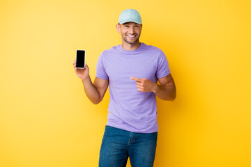 happy man in blue cap pointing with finger at smartphone with blank screen on yellow