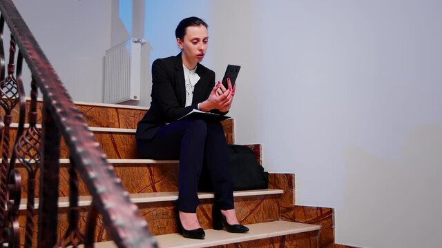 Overworked businesswoman smiling during video call about project deadline sitting on staircase of corporate company building. Entrepreneur talking with client using smartphone on stairs.