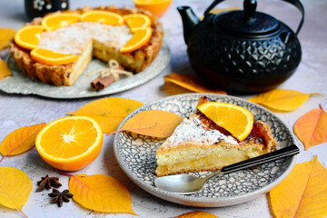 A piece of lemon pie on a plate, oranges, yellow leaves, a teapot on a light gray table. Autumn still life with dlmashny pastries.