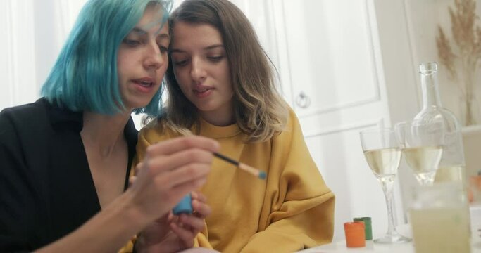 Lesbian couple painting rainbow together. Young girlfriends talking and painting rainbow with gouache together during romantic date with wine at home