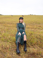 Naklejka na ściany i meble A girl with blonde hair , wearing a black cap, green chiffon dress and rough boots sits in a mown field.