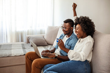 Naklejka na ściany i meble Overjoyed african American couple sit relax on cozy couch look at laptop screen triumph winning lottery online, happy biracial husband and wife feel excited euphoric with good news on computer