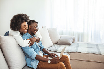 Naklejka na ściany i meble Happy young african couple with laptop computer. Happy black couple working together on laptop sitting on sofa at home. Freelance and remote work concept, copy space