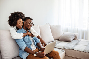 Happy mixed race young couple using laptop looking at screen together watching video movie sit on sofa, smiling interracial husband and wife make online computer call do internet shopping at home