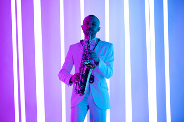 Happy saxophonist plays music on sax in elegant white suit. Man stand in the studio against the...