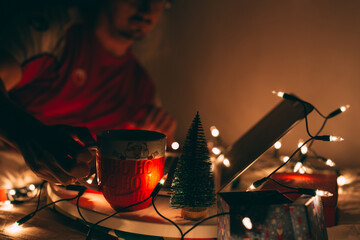 Man using  laptop in bed and having some drink from big mug on Christmas eve, beautiful warm ambient