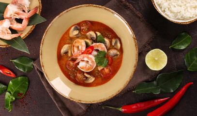 Thai cuisine. Tom yum. Coconut milk soup with shrimp, mushrooms, tomatoes, galangal root, lemongrass, chili pepper in a beige bowl on a brown table. Rice, lime. Background image, copy space, top view