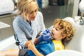 Cute young boy visiting dentist, having his teeth checked by female dentist in dental office.