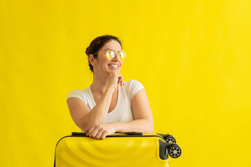 A woman with a perfect snow-white smile in sunglasses hugs a suitcase on a yellow background. Excited girl waiting for travel on summer vacation and snuggles up to her bag.