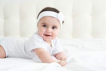 Cute healthy baby girl 6 months smiling in a white bodysuit lying on a bed on white bedding.
