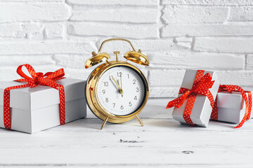 Golden alarm clock and gift boxes with red ribbon on a white wooden table, brick backdrop. Selective focus. Copy space.
