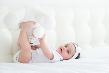 Obraz na płótnie Canvas funny 6 months baby girl playing with legs, while lying in bed at home on white bedding.