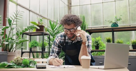 Happy Caucasian businessman talking on cellphone while standing in apron in small floral center and writing down order details. Joyful male florist calling on smartphone at work. Own business concept - Powered by Adobe