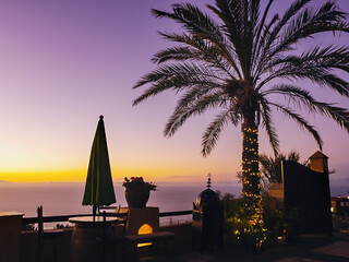 Purple golden evening atmosphere on the north coast of Tenerife, with a large palm tree surrounded by light chain on a viewing terrace overlooking the Atlantic Ocean. A closed parasol and an old woode