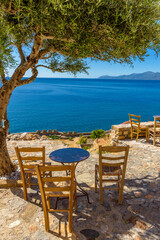 Fototapeta na wymiar Traditional cafe exterior in the fortified medieval castle of Monemvasia. Iron tables and wooden chairs and an olive tree with the view of the aegean sea in the background.