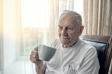 old man is sitting in a chair. A smiling old man is sitting in a chair by the window with a Cup of...