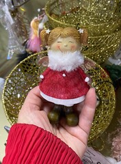 A smiling porcelain doll in red and white winter clothes stands in a human hand against a background of Golden baskets.
