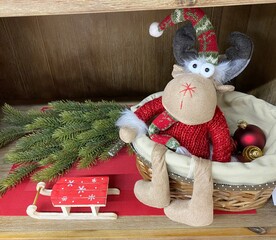 A stuffed Christmas reindeer in red clothes sits in a basket on a shelf next to a toy sleigh and fir branches. 