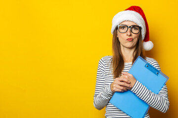 Young woman with a pensive face in a Santa Claus hat holds a clipboard in her hands on a yellow background. Christmas planning and gifts planning concept. Banner