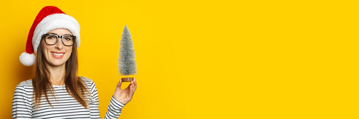 Young woman with a smile in a Christmas hat holds a beautiful Christmas tree on a yellow background. Banner. Christmas and New Year concept