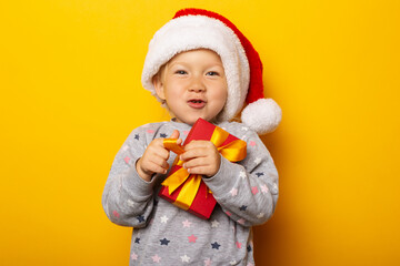 Child in a Santa Claus hat holds a gift on a yellow background. Gifts for Christmas, Christmas morning. Banner