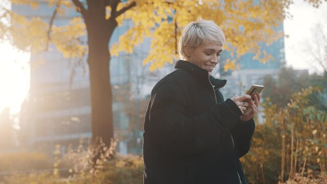 Young caucasian short haired blond woman using phone in the city park in front of office building in autumn. High quality 4k footage