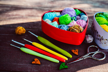 Heart-shaped boxes with multicolored  skeins of yarn for knitting and hooks  on wooden background in the garden on spring day. Shadows. Crochet and knitting. Women's working space.