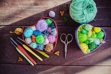 Heart-shaped boxes with multicolored skeins of yarn for knitting and hooks on wooden background in the garden on spring day. Shadows. Crochet and knitting. Women's working space.	