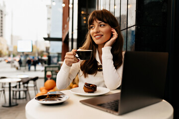 Elegant stylish woman with long dark hair wearing white shirt drinking coffee while working on laptop in outside cafe on background of Christmas lights