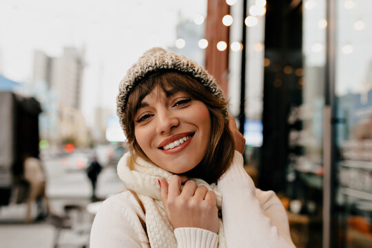 Blissful girl in stylish winter outfit posing in outdoor cafe with modern exterior. Photo of merry caucasian woman enjoying Holiday
