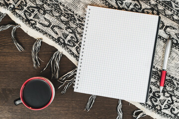 Blank notebook with a pen, a glass of coffee and a woolen warm tablecloth on a wooden table. View from above.