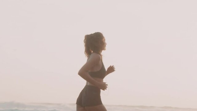 Full shot of toned Caucasian young woman wearing shorts, sports bra an smartphone in armband case jogging alongside oceanfront on sandy beach under sunshine