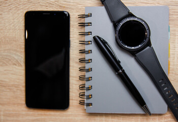 Smartphone and smartwatch next to a notebook on a wooden desk. There is also pen on the desk.