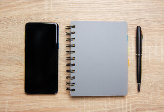 Smartphone next to a notebook on a wooden desk. There is also pen on the desk.