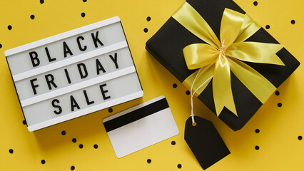 Banner concept for Black Friday celebration,lettering.White board with text BLACK FRIDAY SALE,black gift box with yellow bow,tag,credit card,confetti on yellow background,top view,flat lay,close-up.