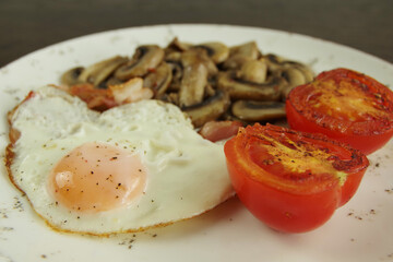 Fried egg with bacon, fried mushrooms,  and tomatoes on a white plate. Breakfast recipe.