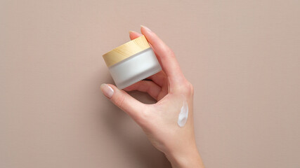 Female hand holding jar of cosmetic cream on pastel beige background. Woman applying moisturizer...