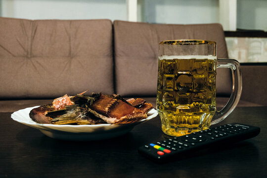 Glass half-liter mug filled with beer, dried smoked fish and TV remote control on the table near the sofa.