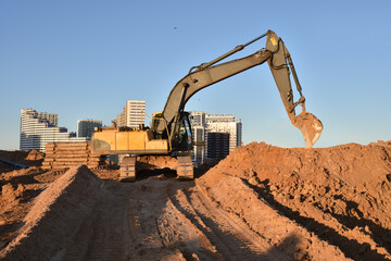 Obraz premium Excavator on earthworks at construction site. Backhoe on foundation work and road construction. Tower cranes in action on blue sky background. Heavy machinery and construction equipment