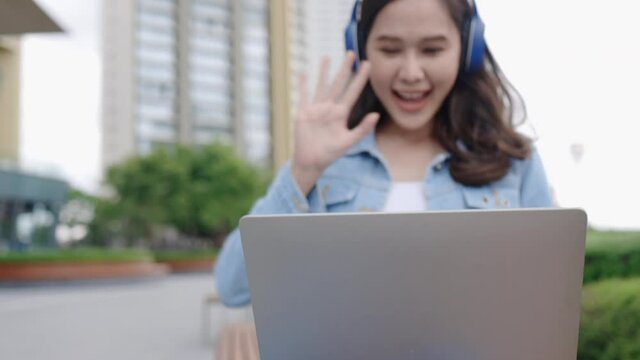 Happy Young Asian women Use the laptop and Headphones to Education or work in the city park. Meeting or study outdoors using free Wi-Fi internet access. Concept technology video conference
