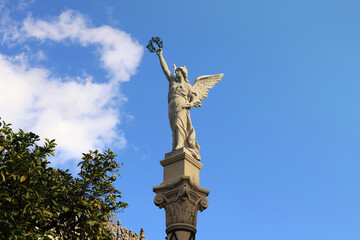 Statue of a female angel with wings and a laurel wreath in the hand, with a blue sky in the...
