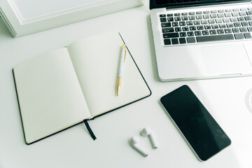 Minimal working desk in office with blank book, smartphone, laptop, and earphones.